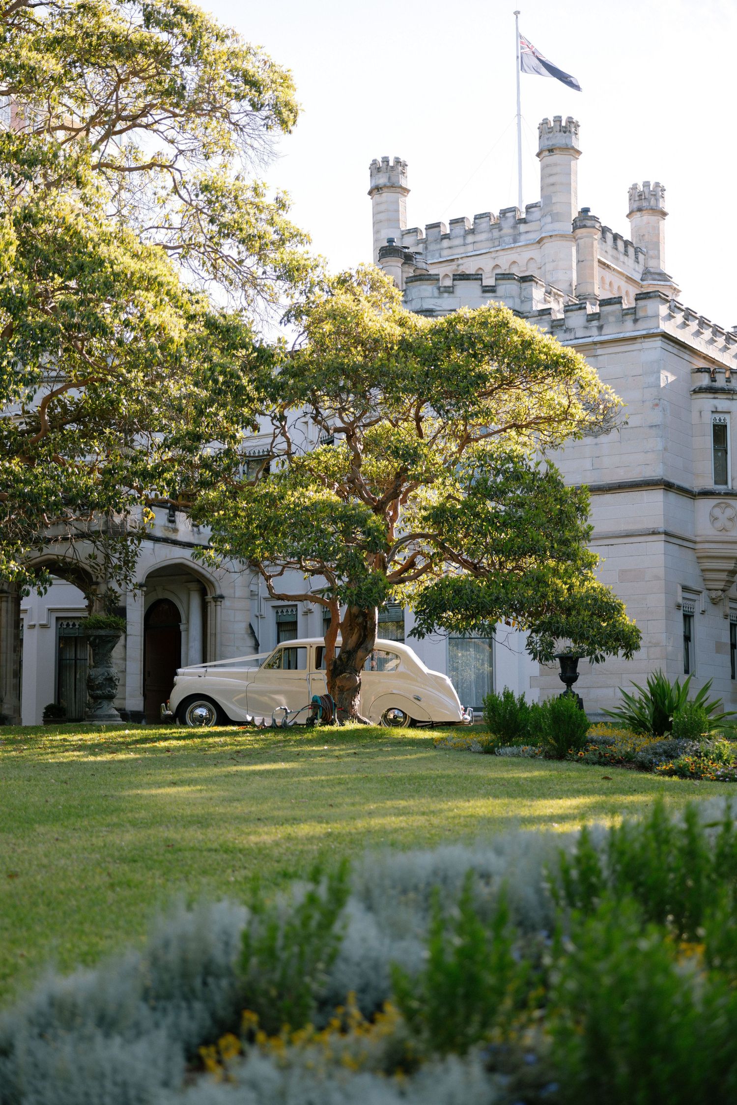 Vintage cream classic car parked on manicured lawn before Gothic castle wedding venue at golden hour.