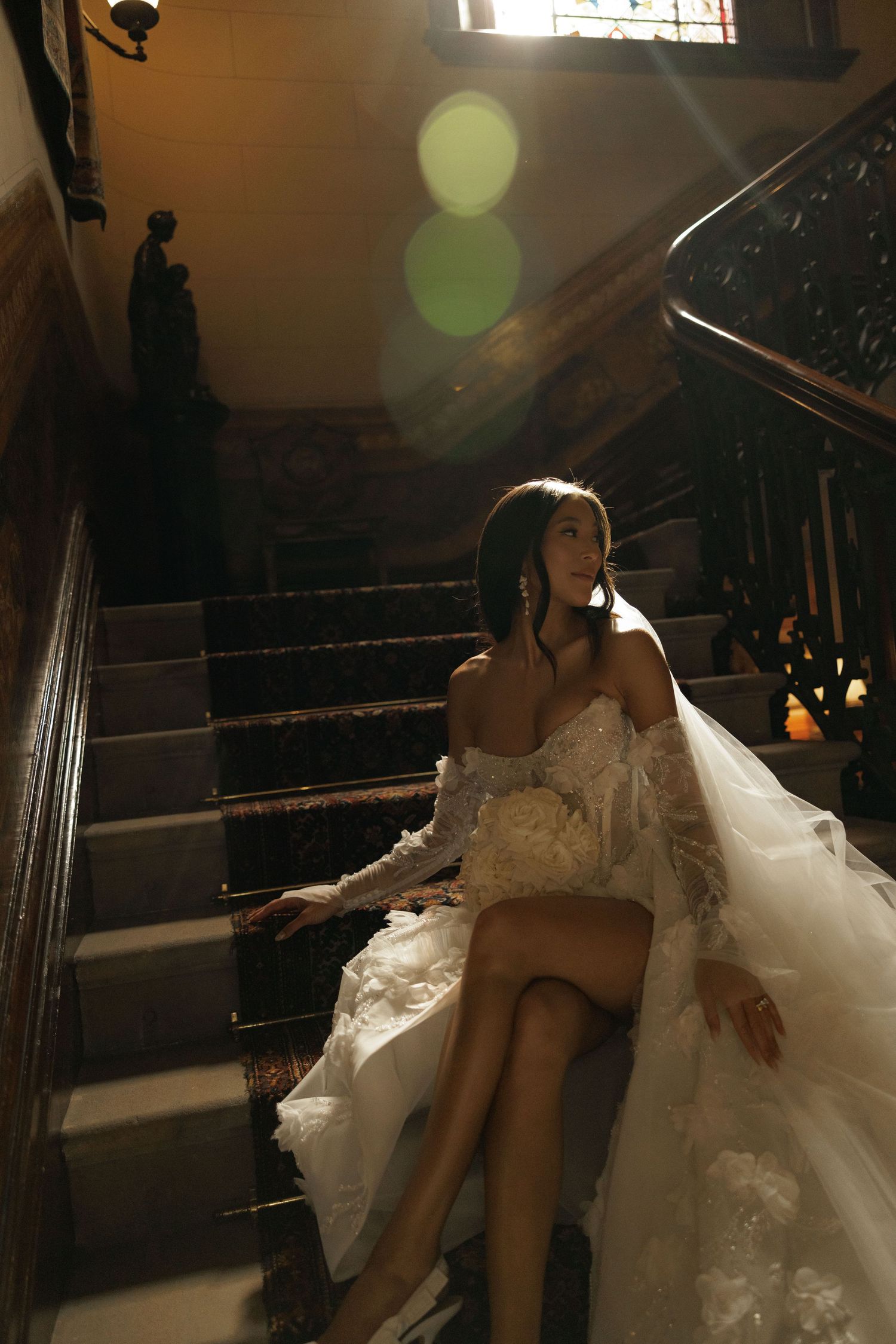 Bride in off-shoulder floral gown sits on grand staircase with cathedral veil, bathed in golden sunlight.