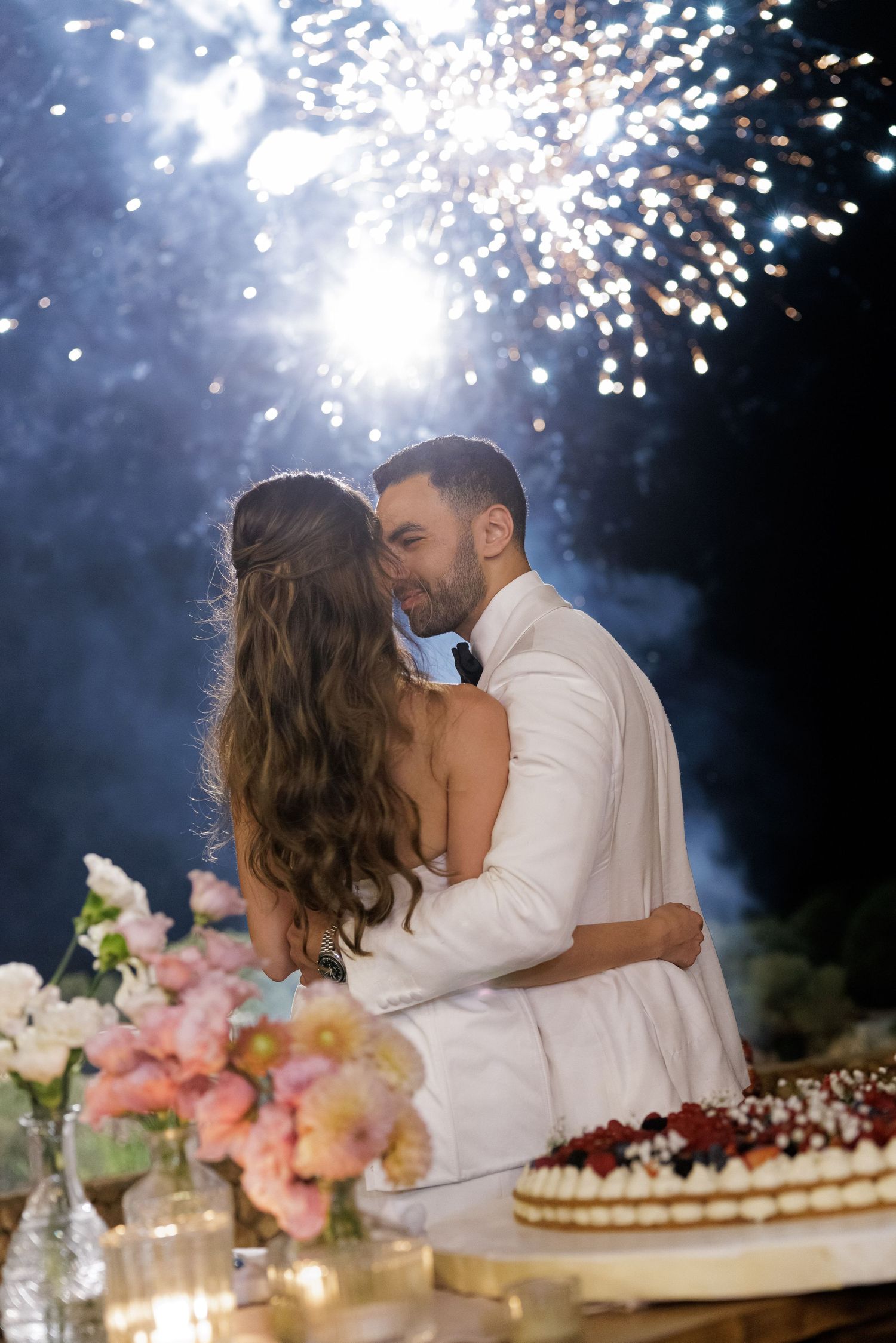 Newlyweds share a romantic kiss beside wedding cake with fireworks display behind them.