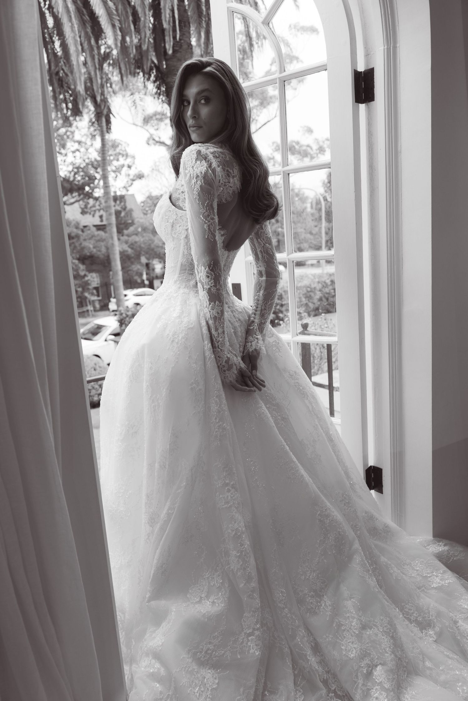 Black and white photo of bride in lace ball gown with long train standing by arched window.