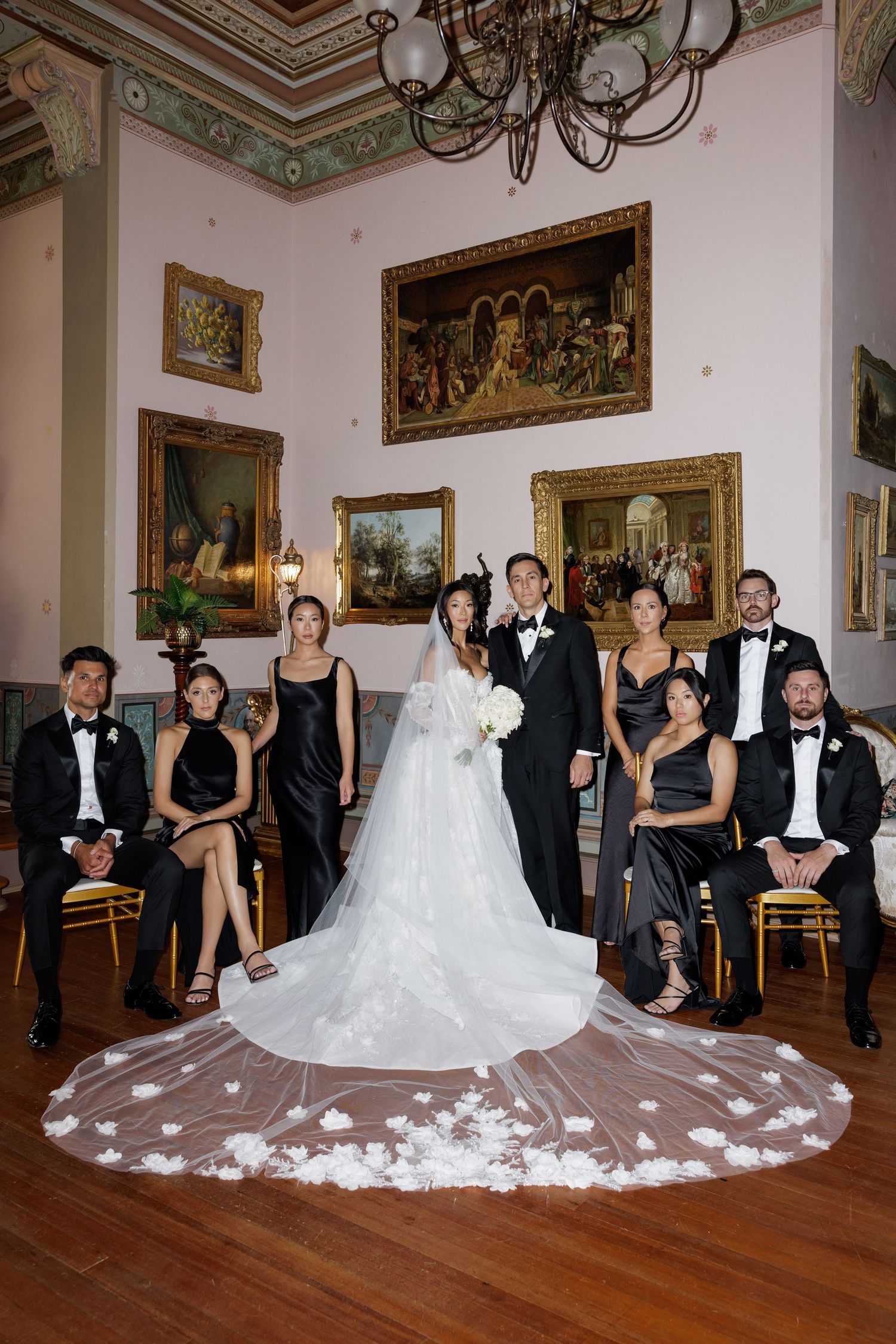 Wedding party portrait in grand room with bride in white gown and attendants in black formal wear.
