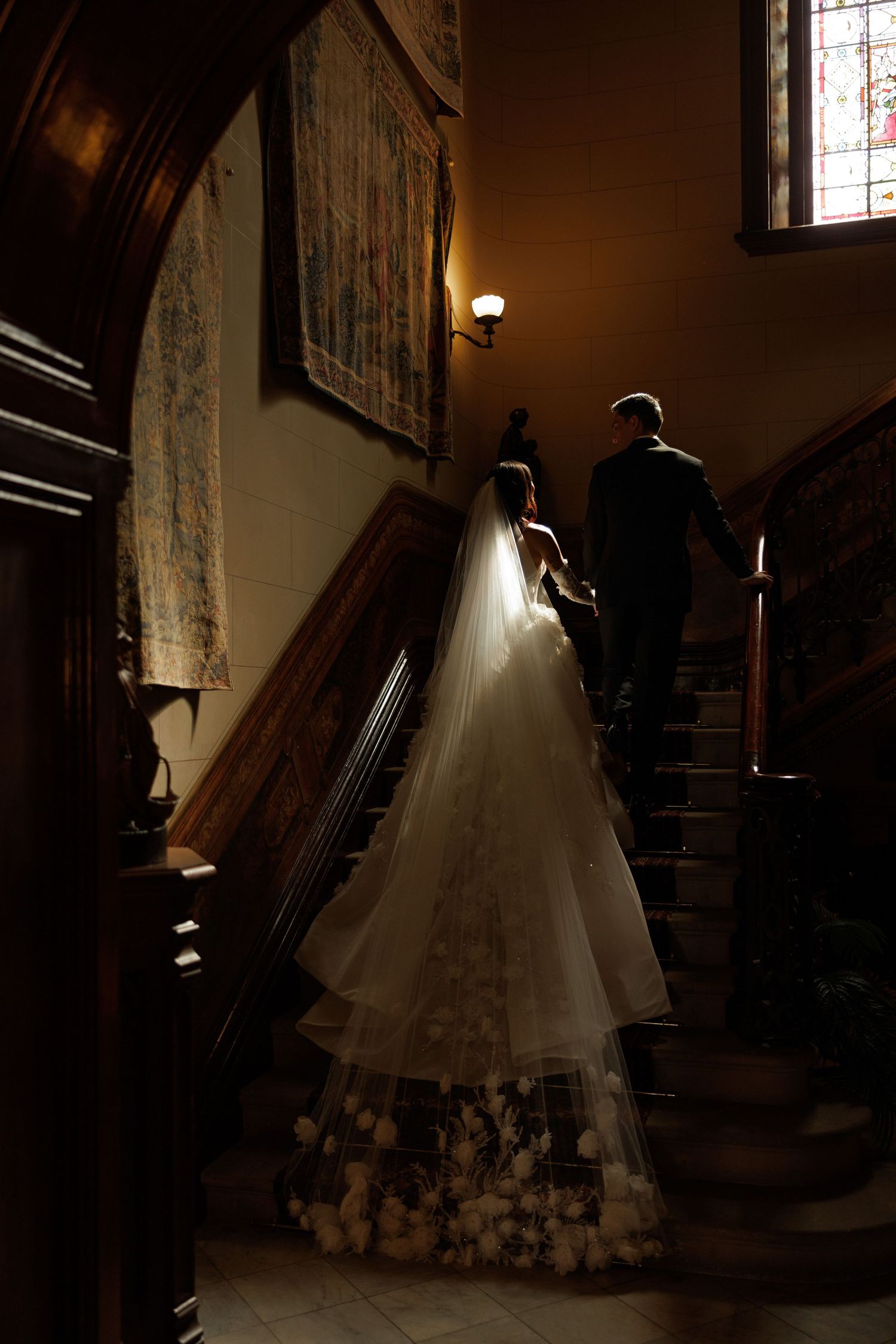Bride in a floral lace gown and long veil walks up a grand dark wood staircase.
