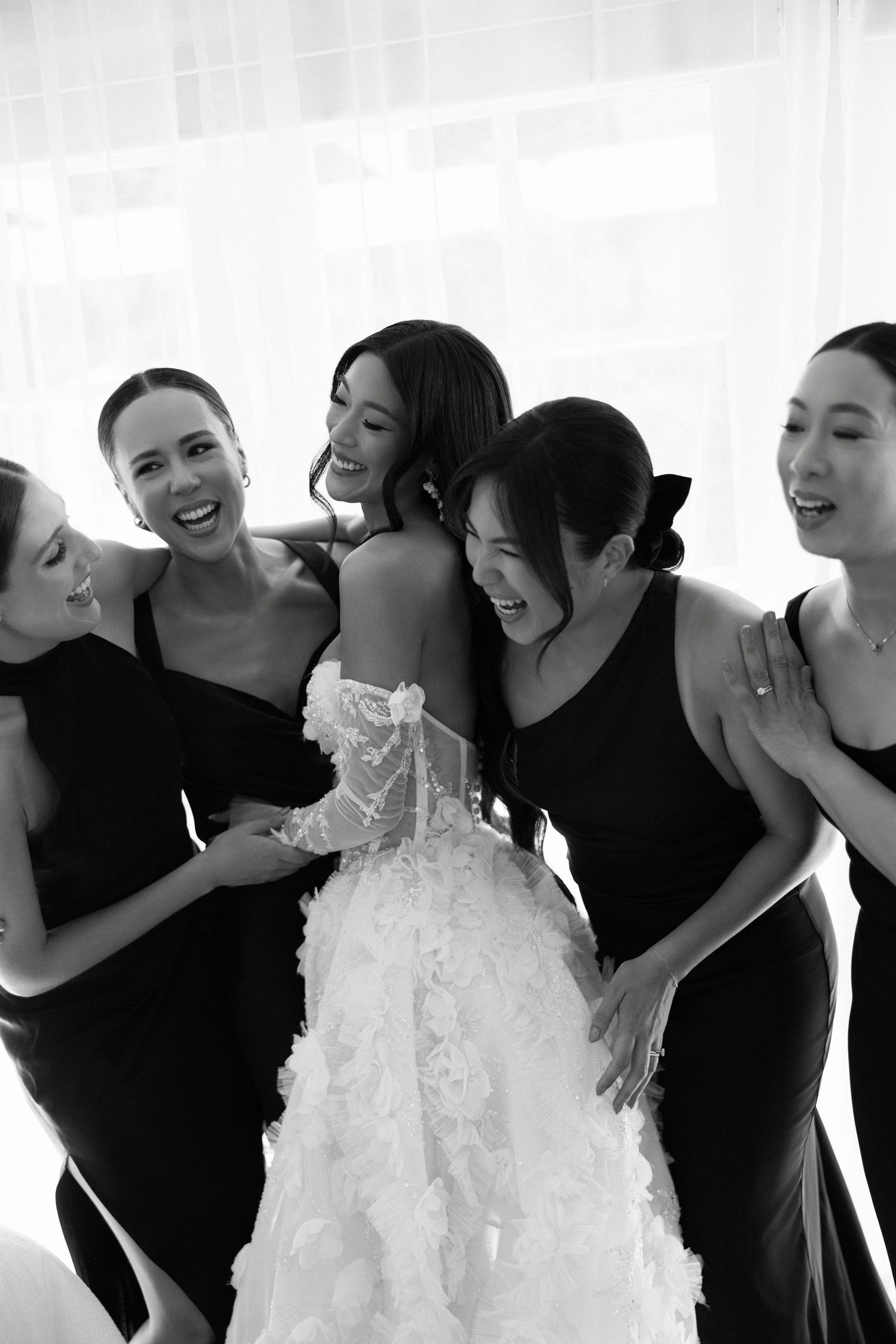 Black and white photo of bridesmaids hugging bride in floral ball gown wedding dress.