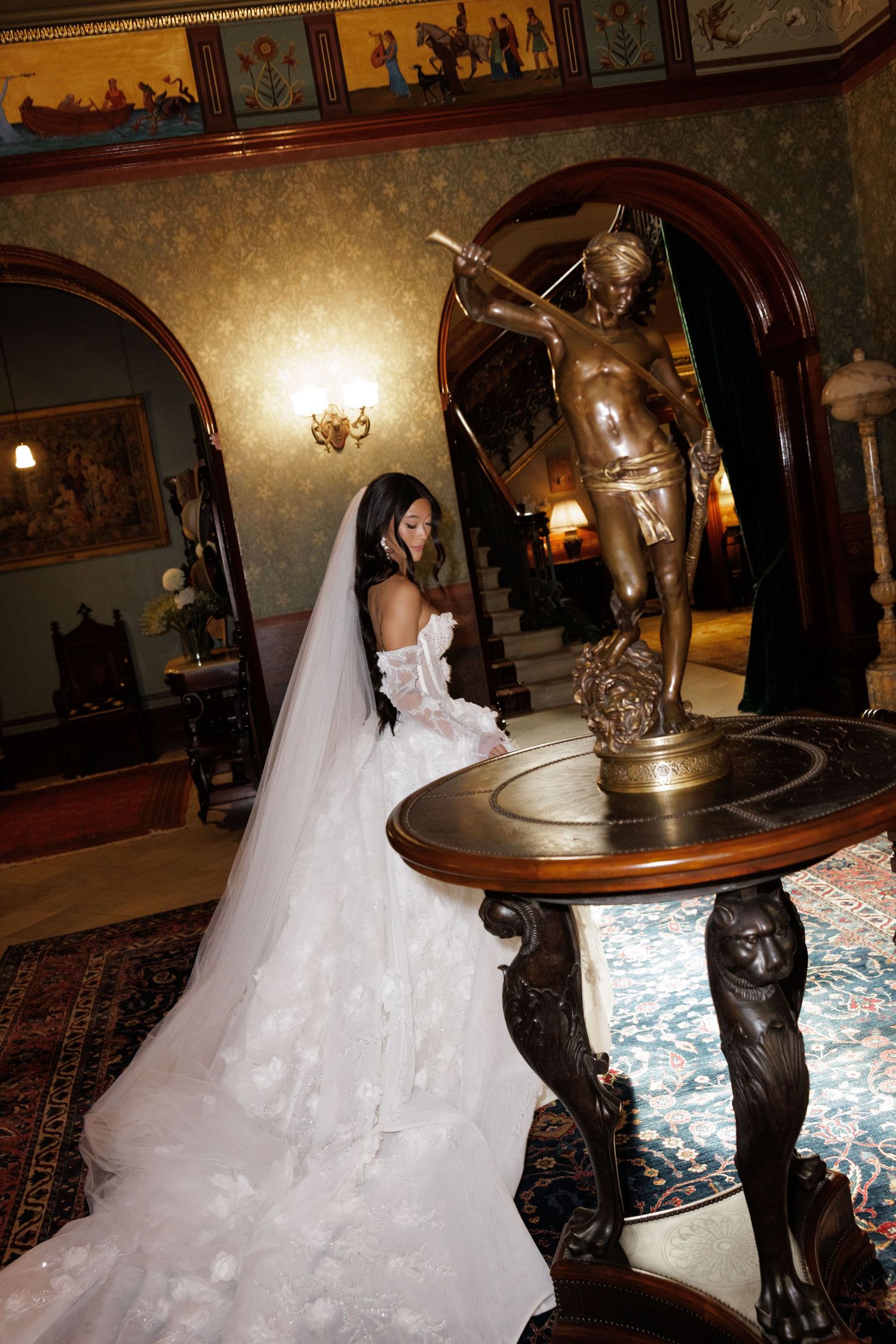 Bride in floral ball gown wedding dress stands beside ornate bronze statue in luxurious mansion interior.