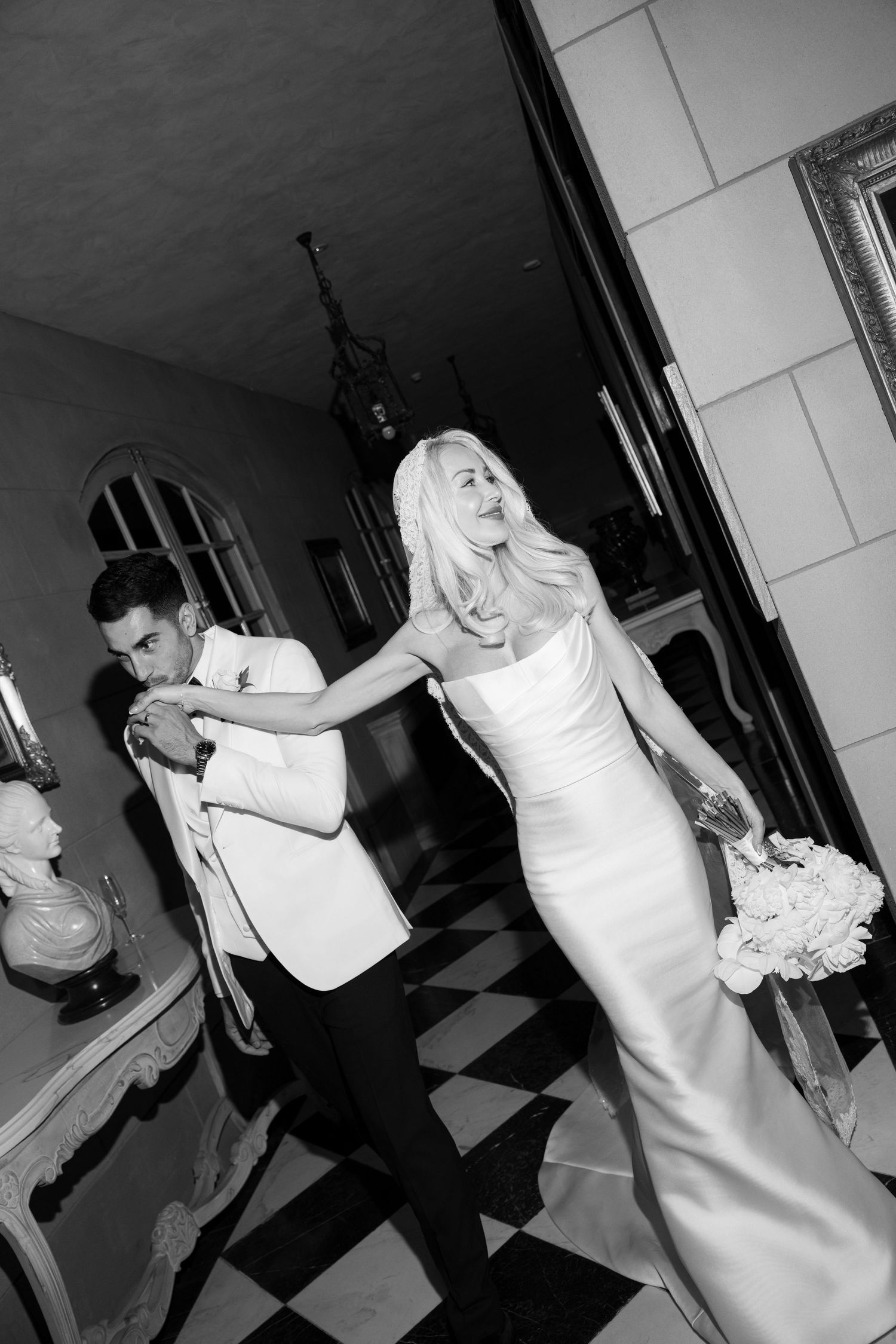 Black and white photo of a bride in a sleek white gown holding flowers, dancing with a groom in a white tuxedo.