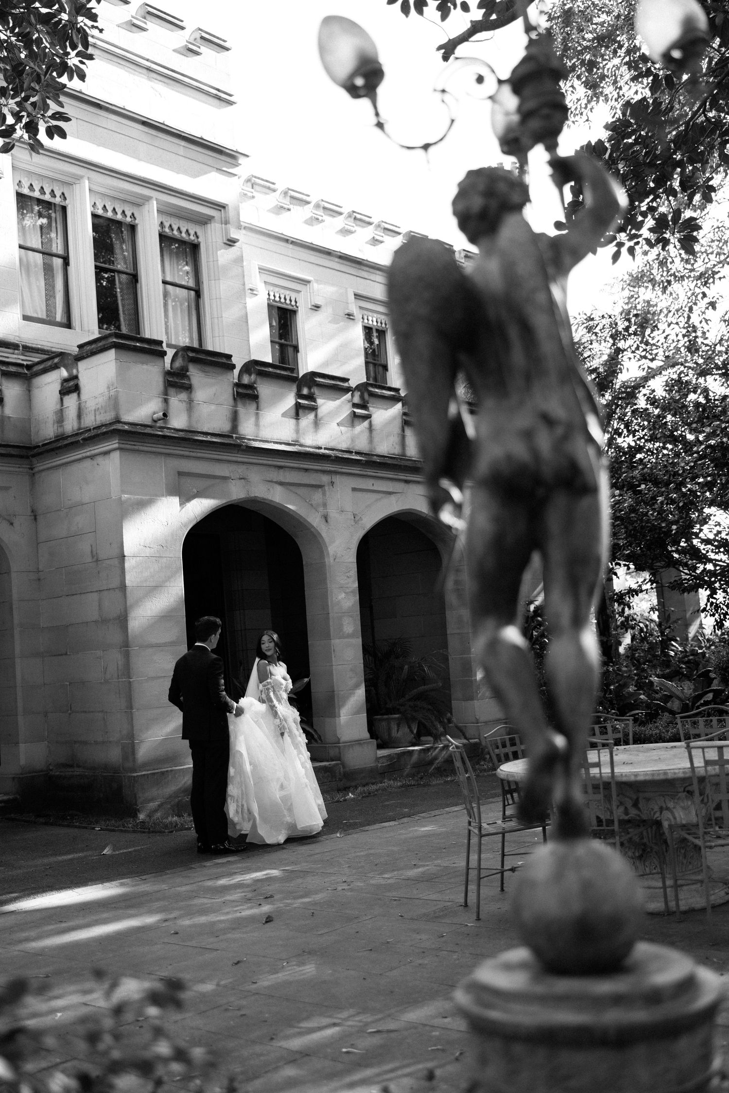 Bride and groom share romantic moment outside grand arched mansion with bronze cherub statue in foreground.