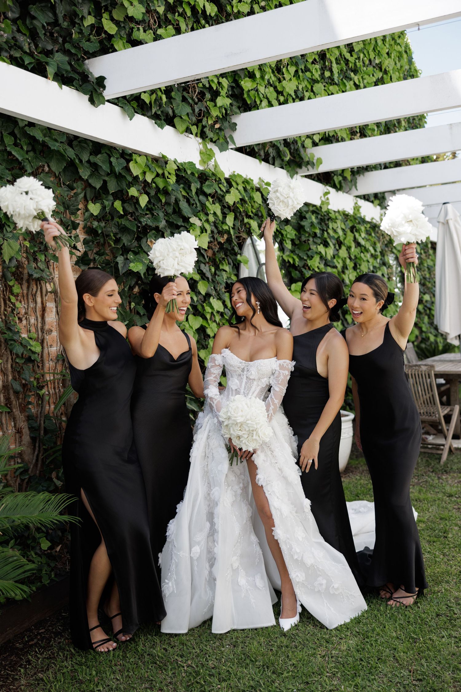 Bride in white strapless gown celebrating with four bridesmaids in black dresses holding white hydrangea bouquets outdoors.