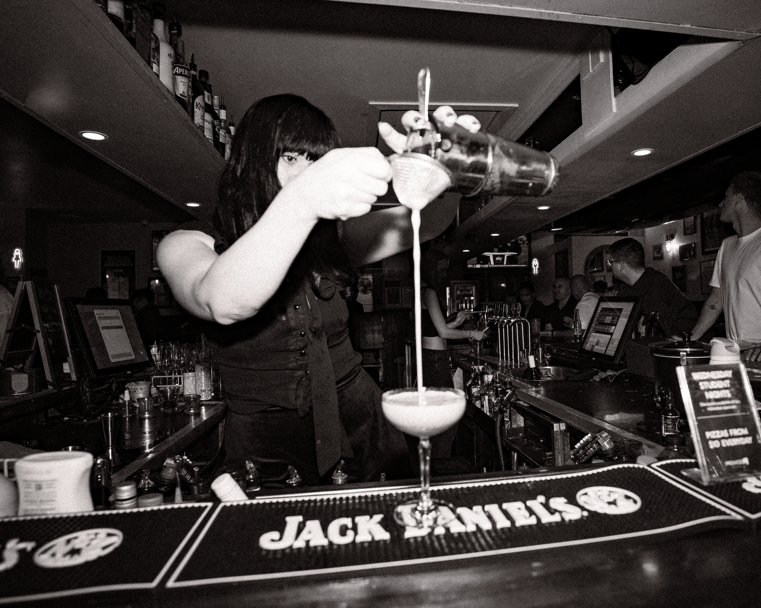 Black and white photo of female bartender pouring a cocktail shaker at a Jack Daniels branded bar.