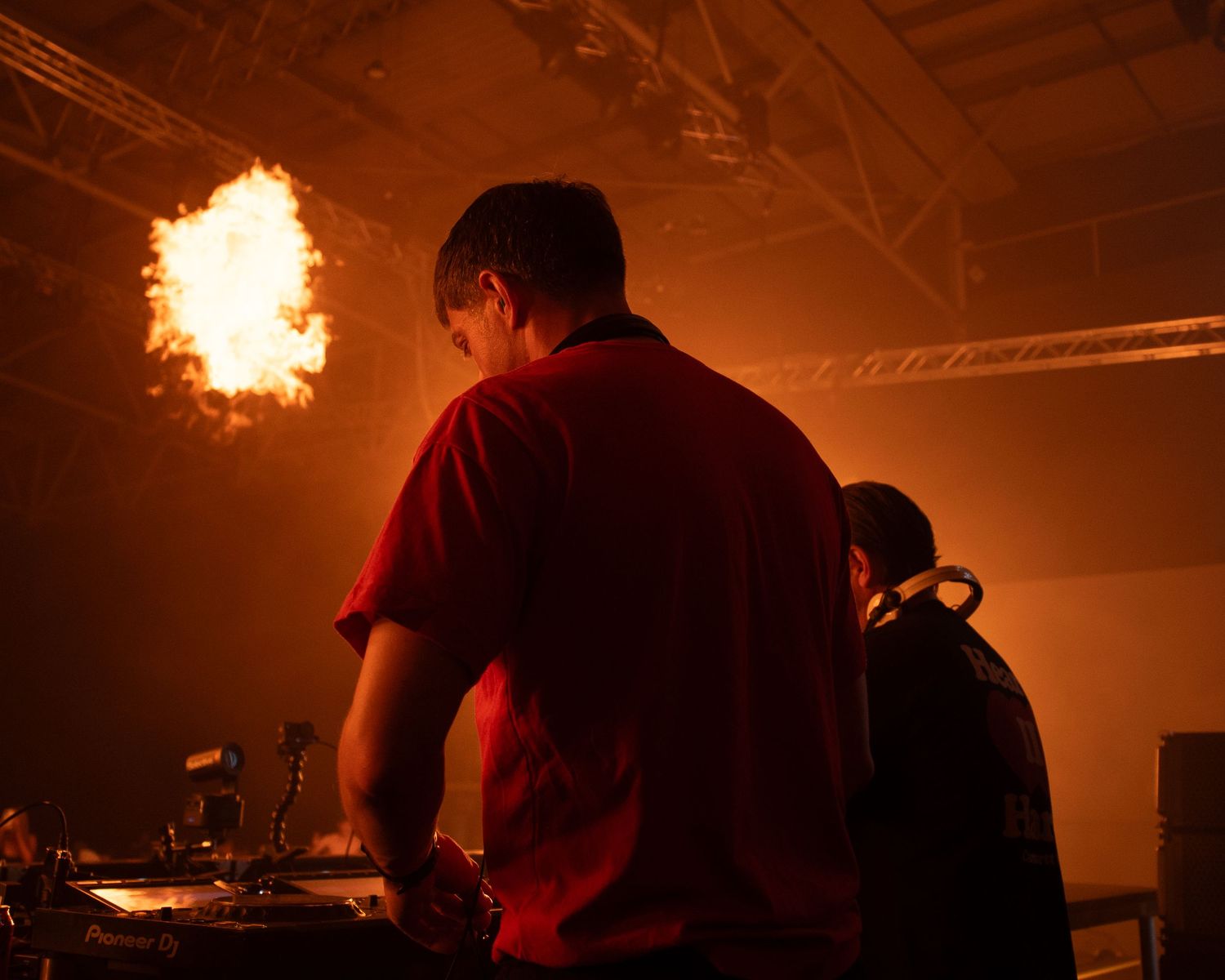 DJ in red shirt performs at a concert with a dramatic fire explosion lighting up the dark venue.