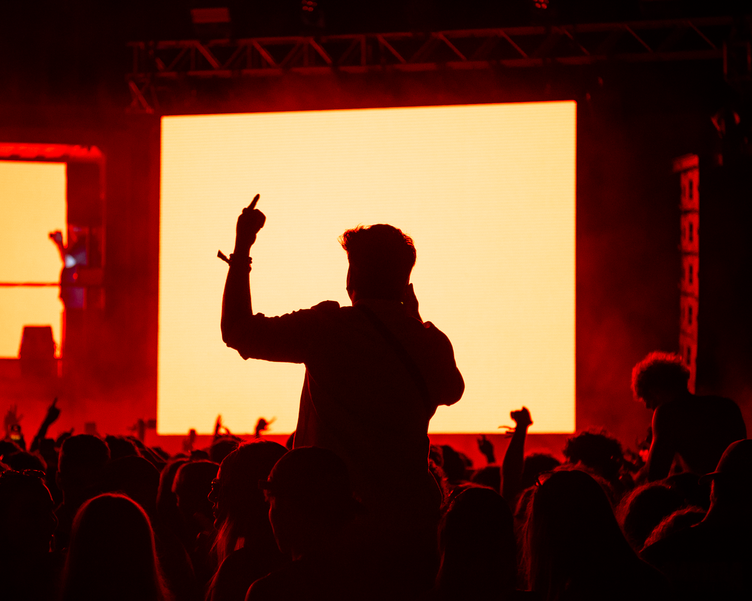 Silhouetted crowd member raised on shoulders gesturing at a glowing yellow screen at a live concert.