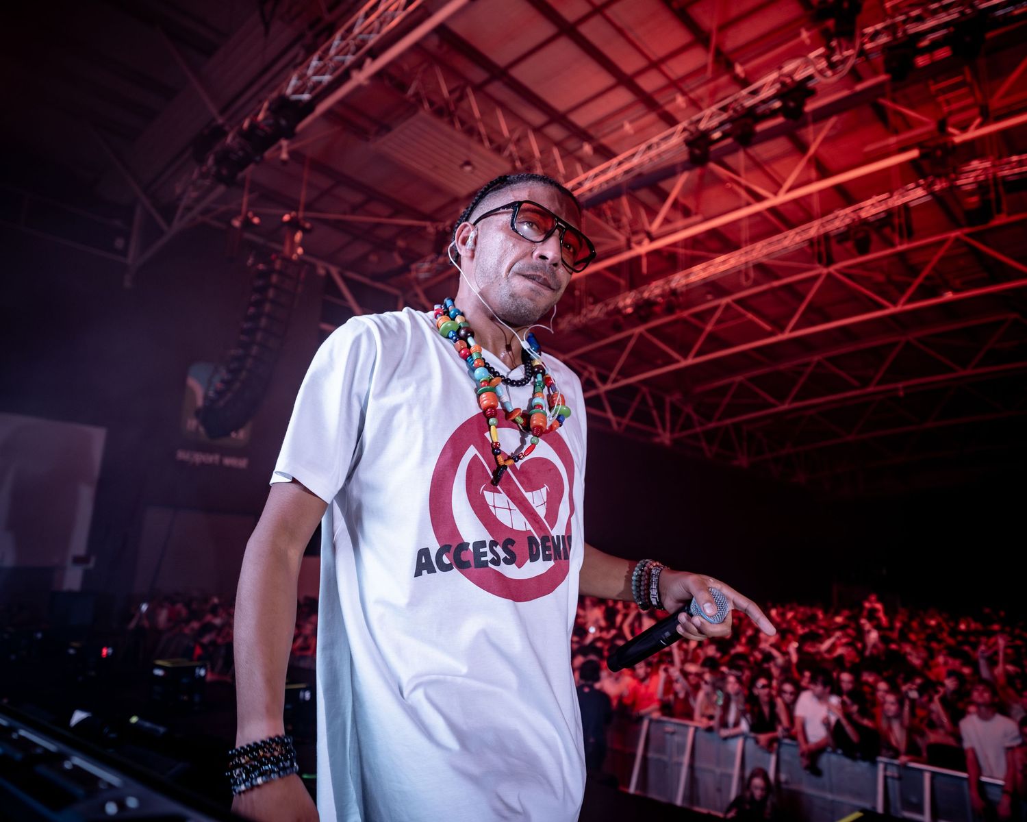 Performer in white Access Denied t-shirt stands on stage before a large crowd at an outdoor music event.