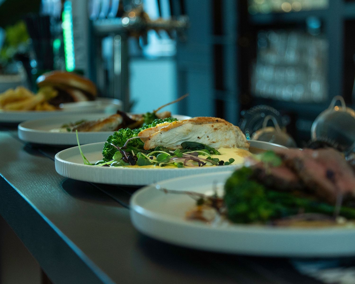 Elegant plated dishes lined up on a restaurant pass, featuring grilled chicken and fresh salads.