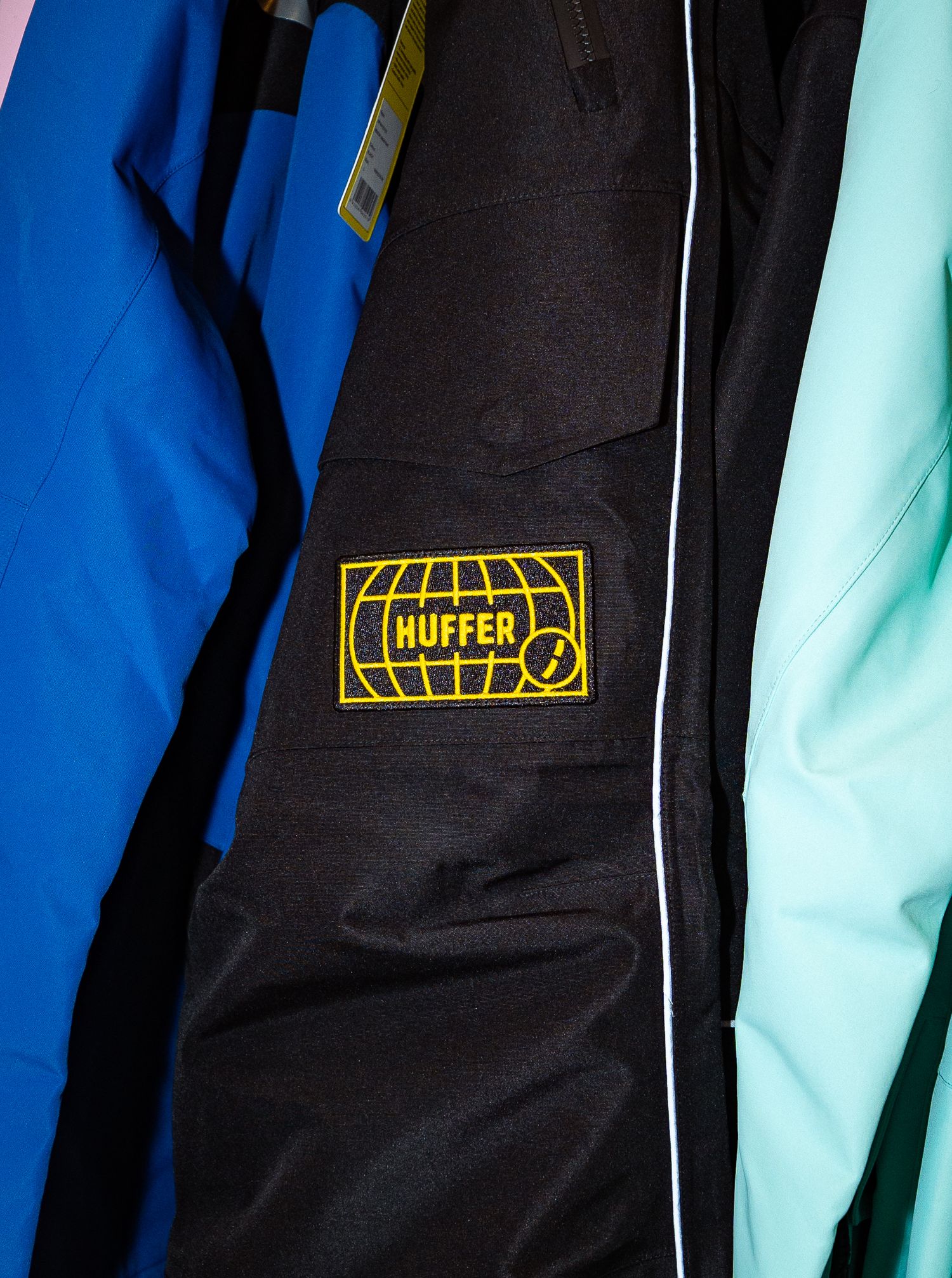 Close-up of a black Huffer jacket with a yellow embroidered logo patch, flanked by blue and mint green jackets.