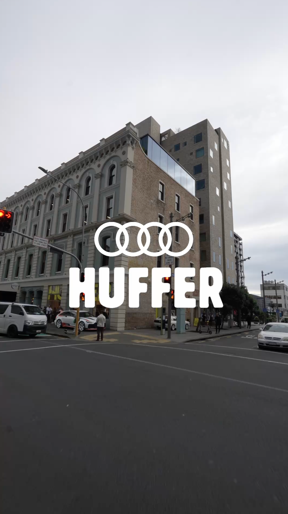 Huffer brand logo overlaid on a street corner with a historic stone building and modern tower in an urban city setting.