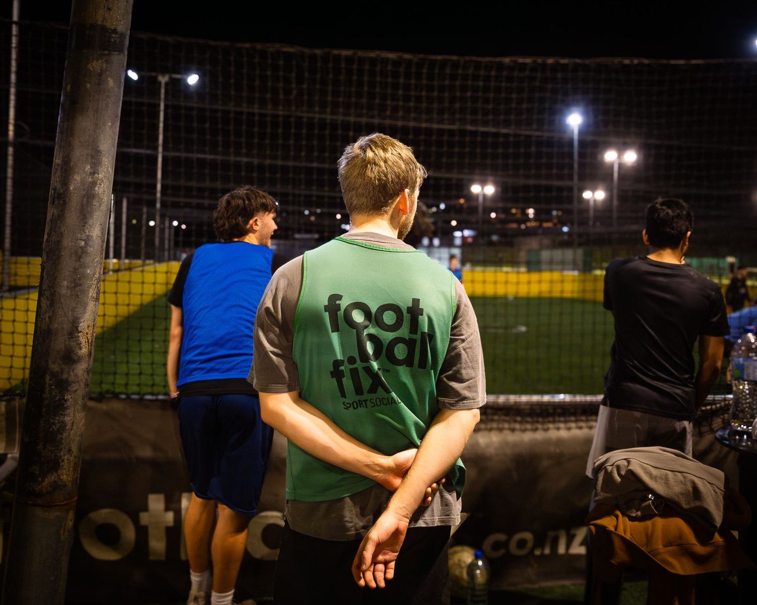 Person wearing a Football Fives green vest watching an indoor football match at night.