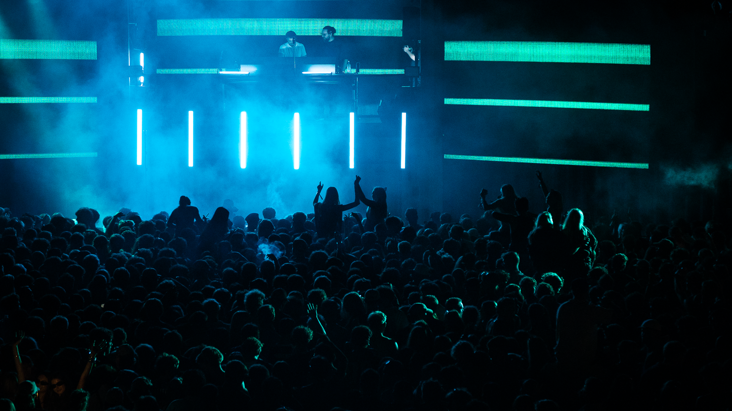 Large crowd of concert attendees bathed in blue-green neon light from vertical LED bars on a dark stage.