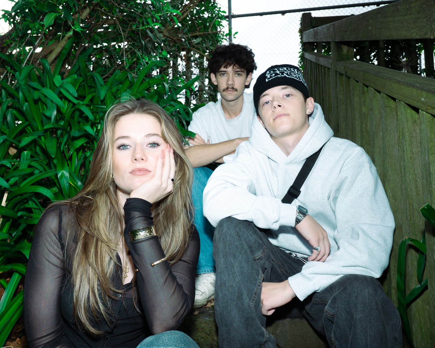 Three young people posing casually on wooden steps surrounded by green garden foliage.
