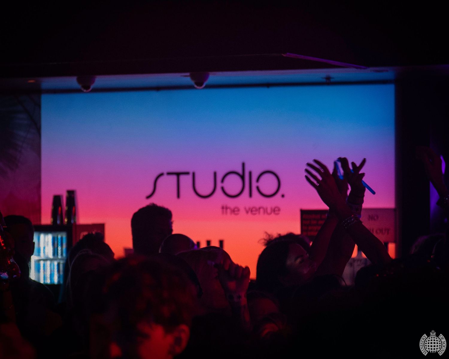 Crowd cheers beneath a colorful Studio The Venue projection screen at a nightclub event.