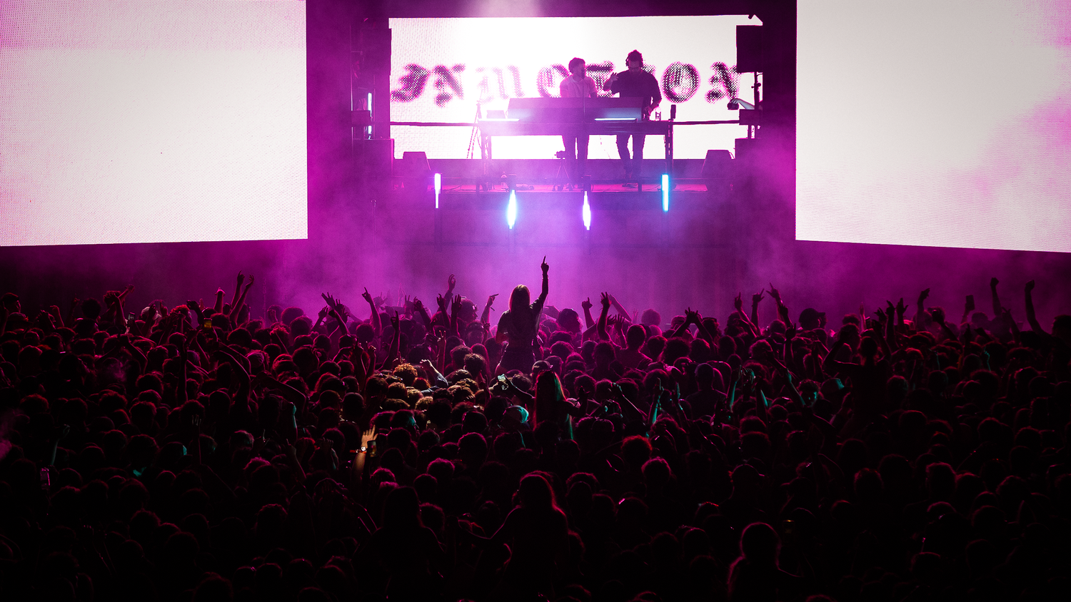 Purple-lit concert stage with performers and large LED screen displaying text before an enthusiastic crowd.