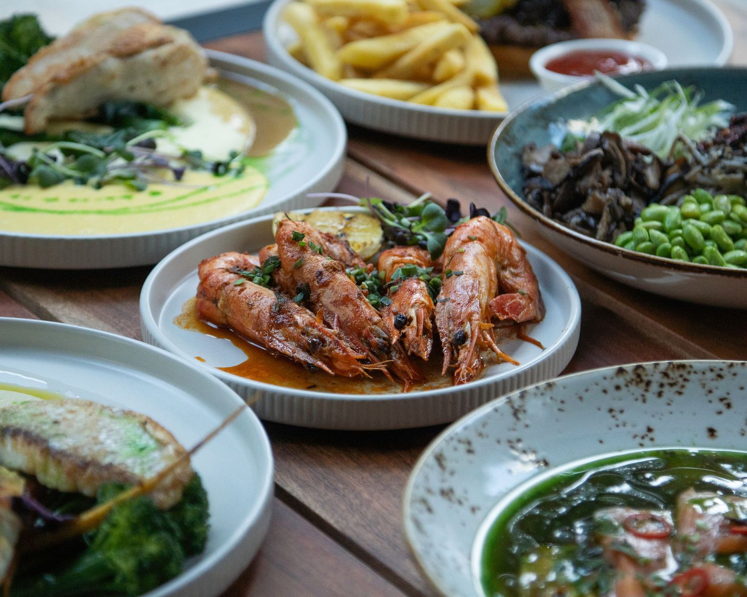 Spread of gourmet dishes including prawns, fries, and greens on a wooden table.