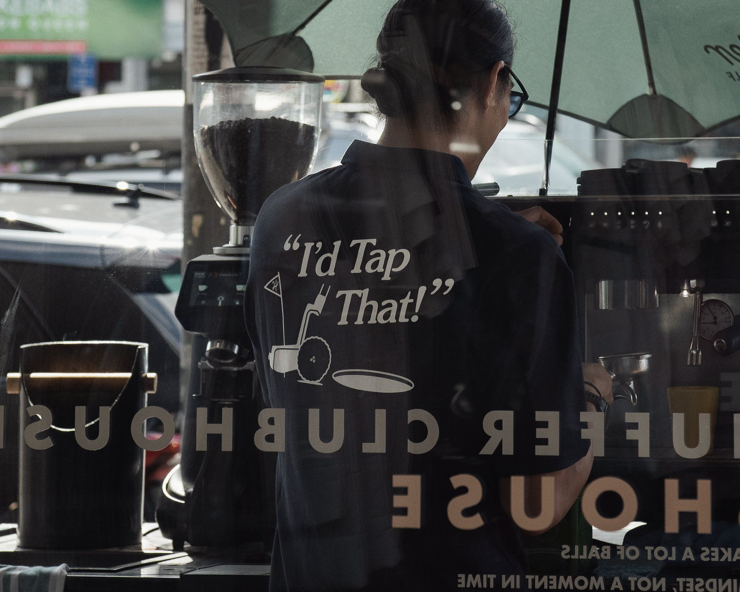Barista wearing I'd Tap That shirt working at Huffer Clubhouse coffee bar reflected in window.