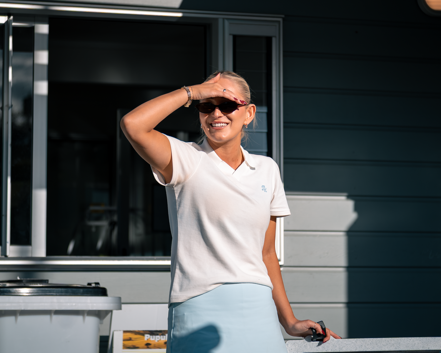 Smiling woman in white polo shirt and light blue skirt shielding eyes from sun outdoors.