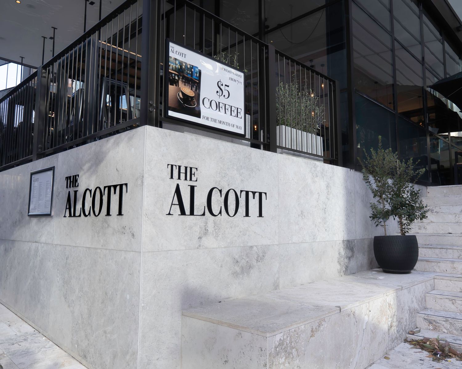 White marble signage of The Alcott restaurant entrance with potted olive tree and steps.