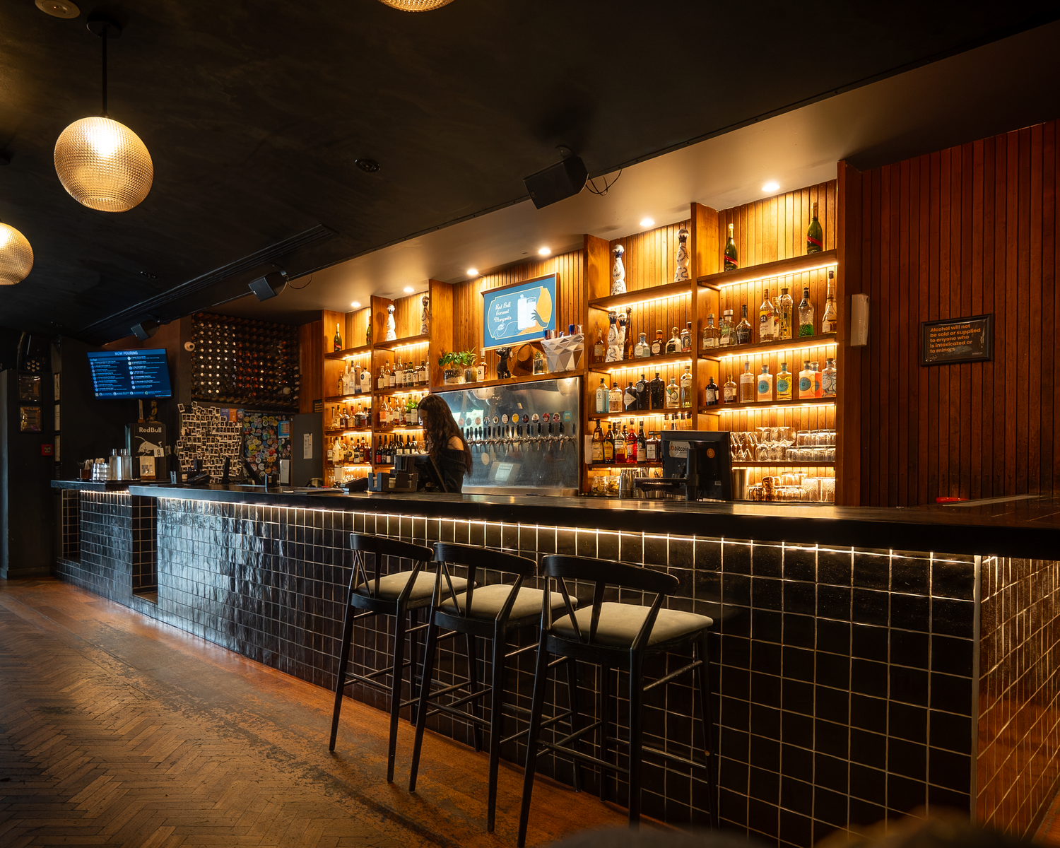 Elegant bar interior with dark tiles, warm lighting, and shelves lined with premium spirits and liquor bottles.