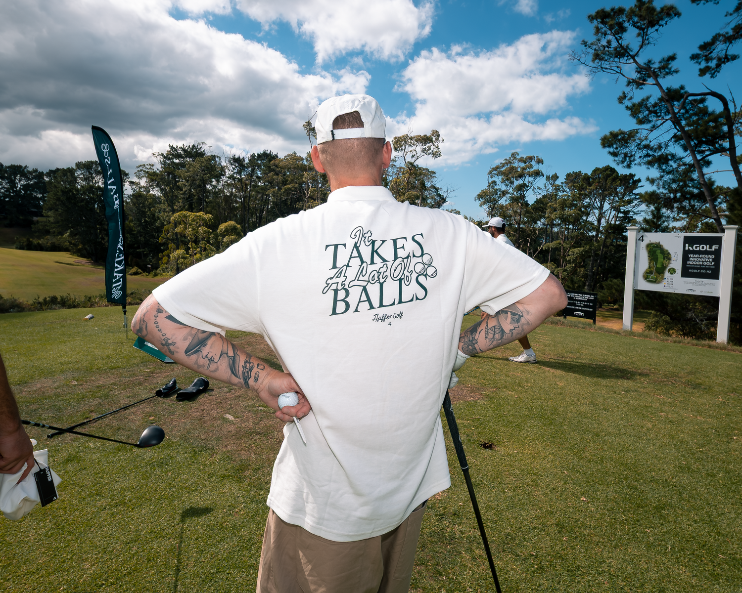 Tattooed golfer in white t-shirt reading Takes Balls on golf course.