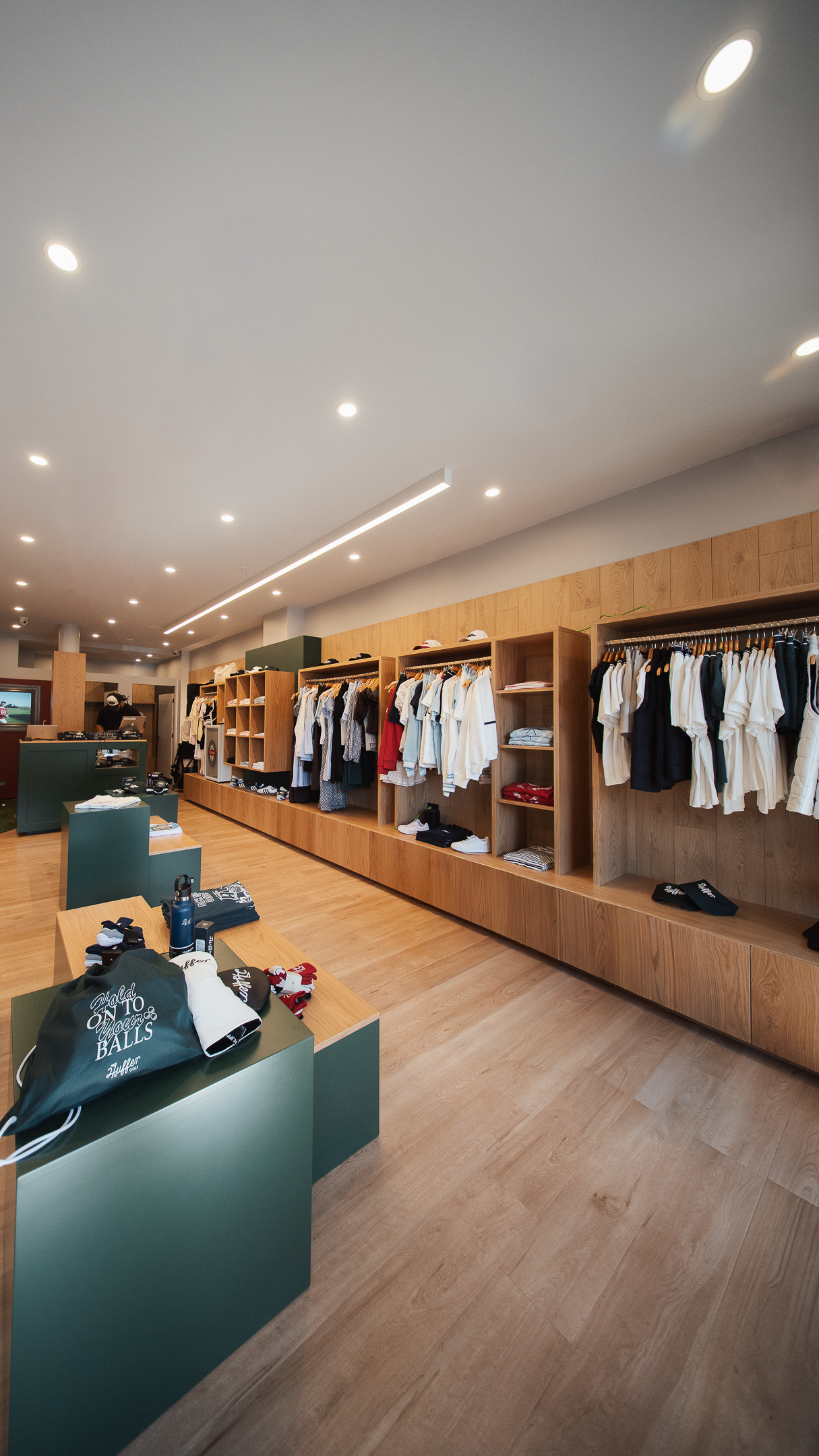 Modern golf apparel retail store interior with wooden shelving, hanging clothes, and green display tables.
