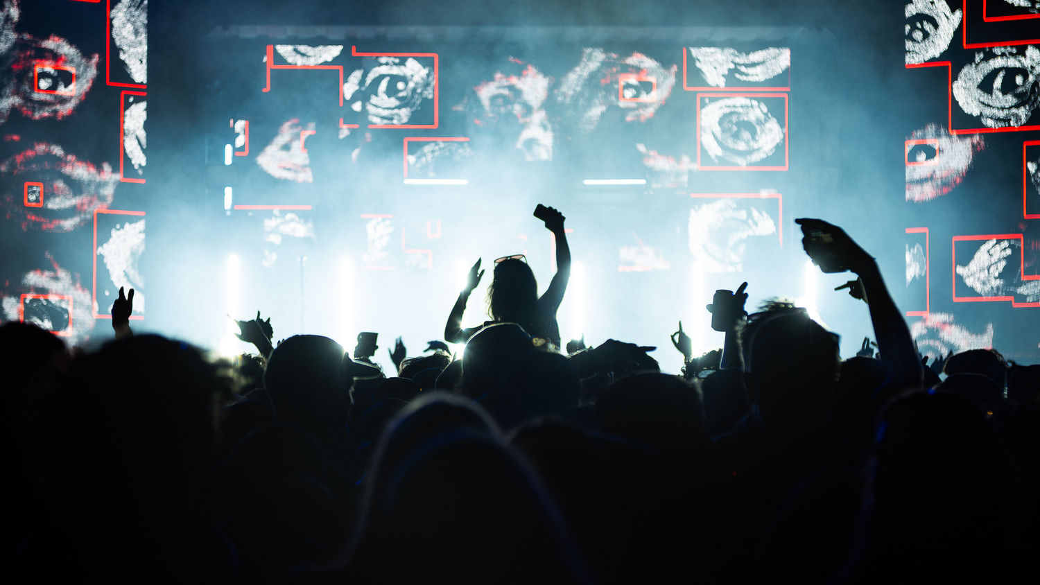 Crowd of silhouetted concert-goers raise hands before a bright stage with large LED screens displaying abstract visuals.