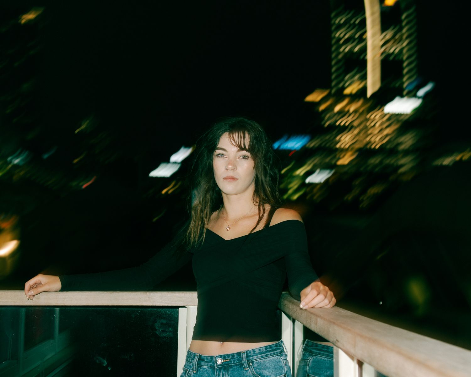 Young woman with dark hair poses on a balcony at night with city lights glowing behind her.