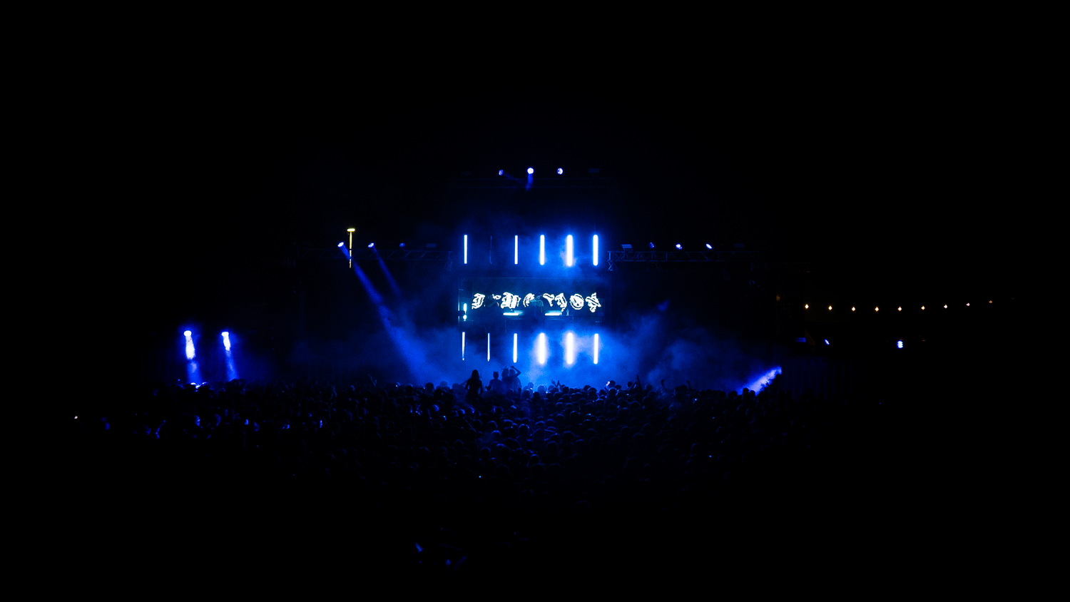 Blue stage lighting illuminates performers at a large outdoor concert in the dark night.