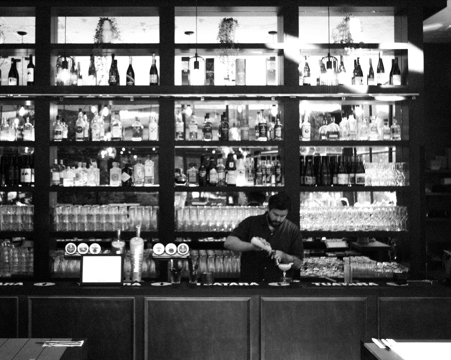 Black and white photo of a bartender working behind a well-stocked bar with bottle displays.