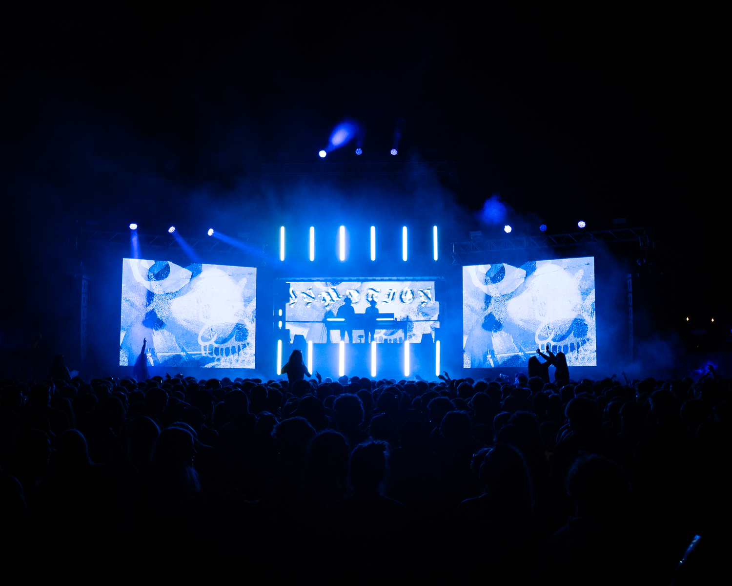 Massive outdoor concert stage with blue LED screens and light beams over a large silhouetted crowd at night.