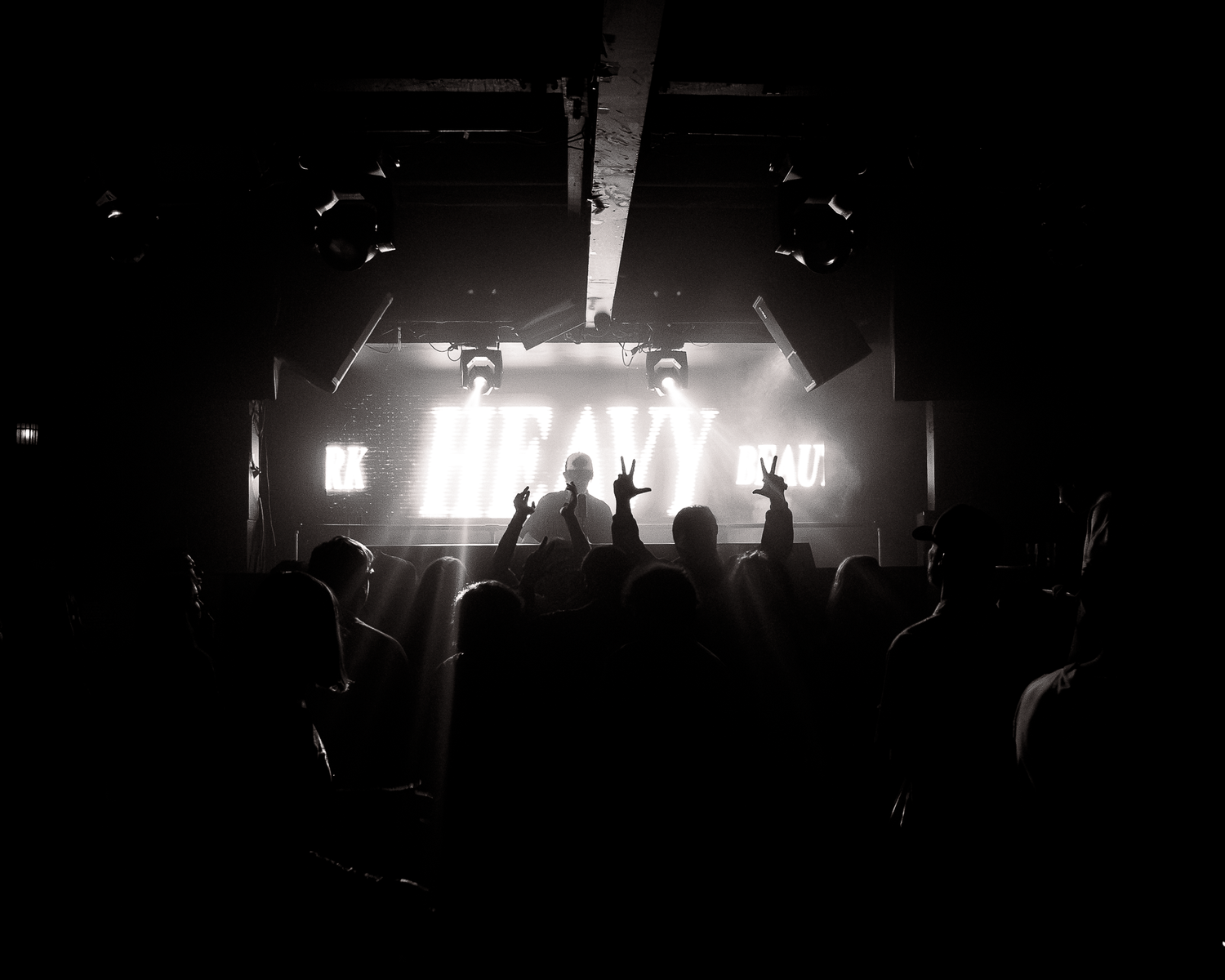 Silhouetted crowd raises hands at a dark concert venue with a bright HEAVY sign on stage.