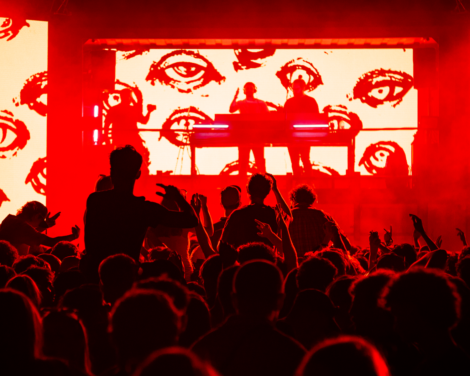 Two performers on stage in red-lit venue with eye-patterned backdrop and cheering crowd below.