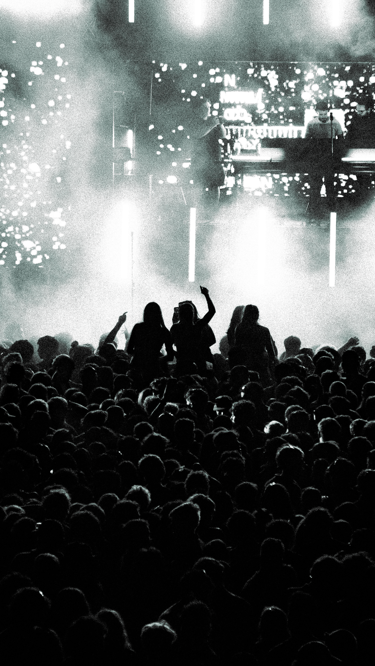 Black and white photo of a massive concert crowd with raised hands and bright stage lights in the background.
