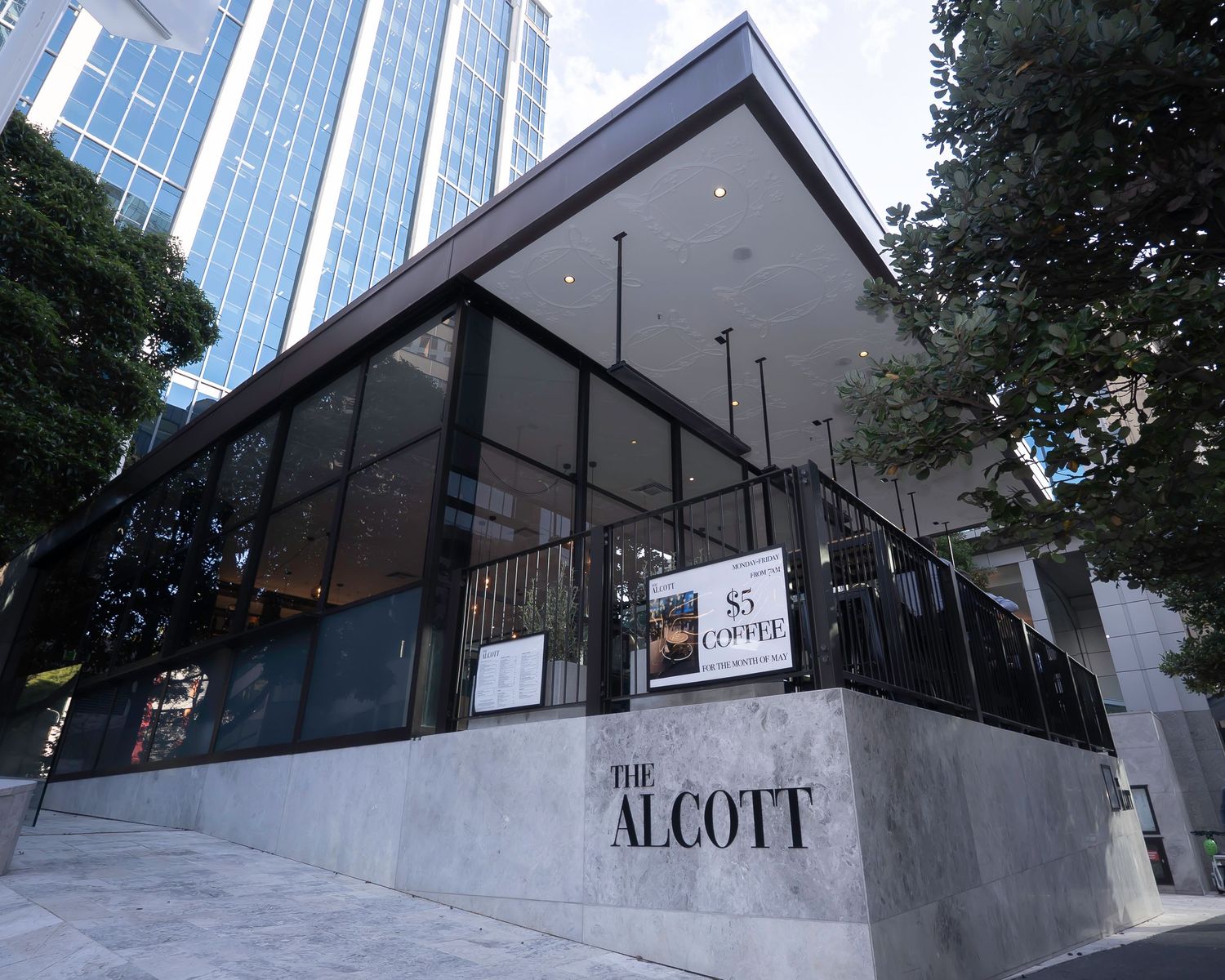 The Alcott restaurant exterior featuring modern black glass facade with 85 Coffee branding and lush trees.