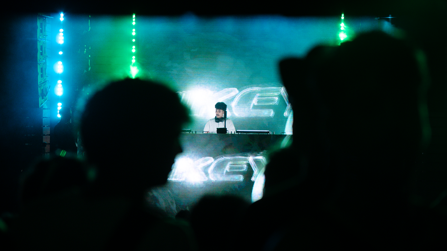 DJ performing on a glowing green and white lit stage with silhouetted crowd in foreground.