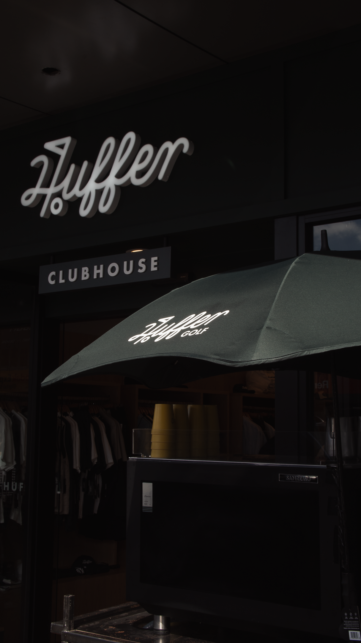 Huffer Clubhouse store exterior with neon sign and branded green umbrella at night.