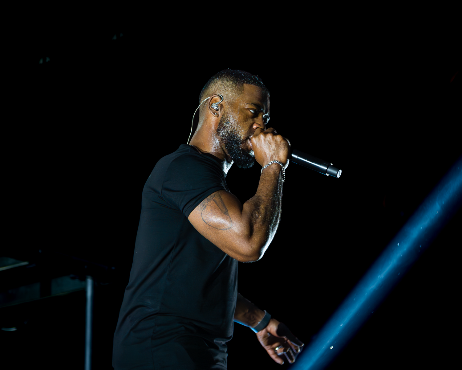 Male performer in black t-shirt singing into microphone on dark concert stage under blue light.