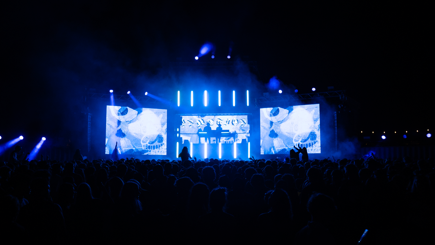 Concert stage lit in blue with large LED screens and performers silhouetted before an energetic crowd.