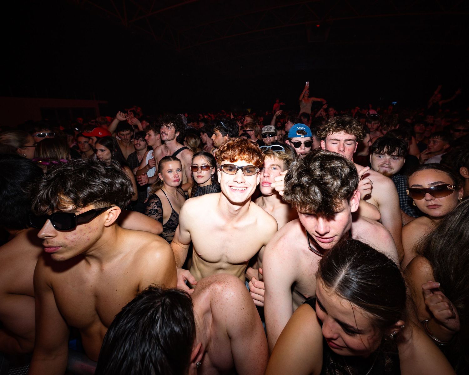 Large crowd of young people at an outdoor night party, many shirtless, packed together under dark sky.