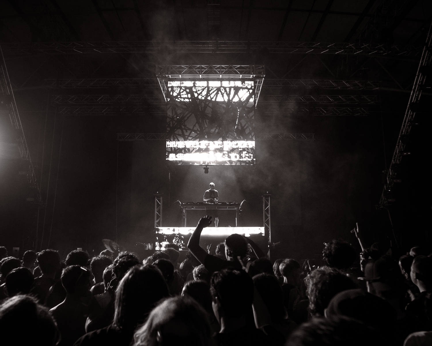 Black and white photo of a DJ performing on stage before a large crowd at a music festival.