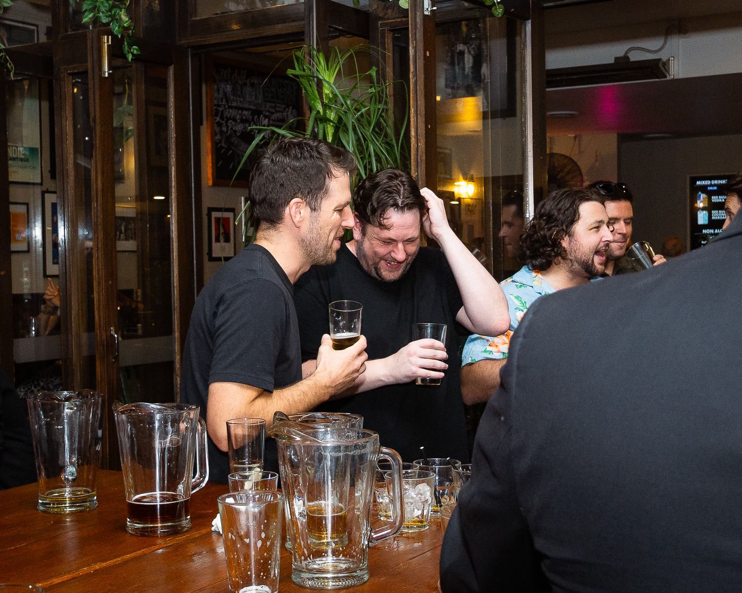 Two men in black shirts laughing and drinking beer at a bar with glass mugs on counter.