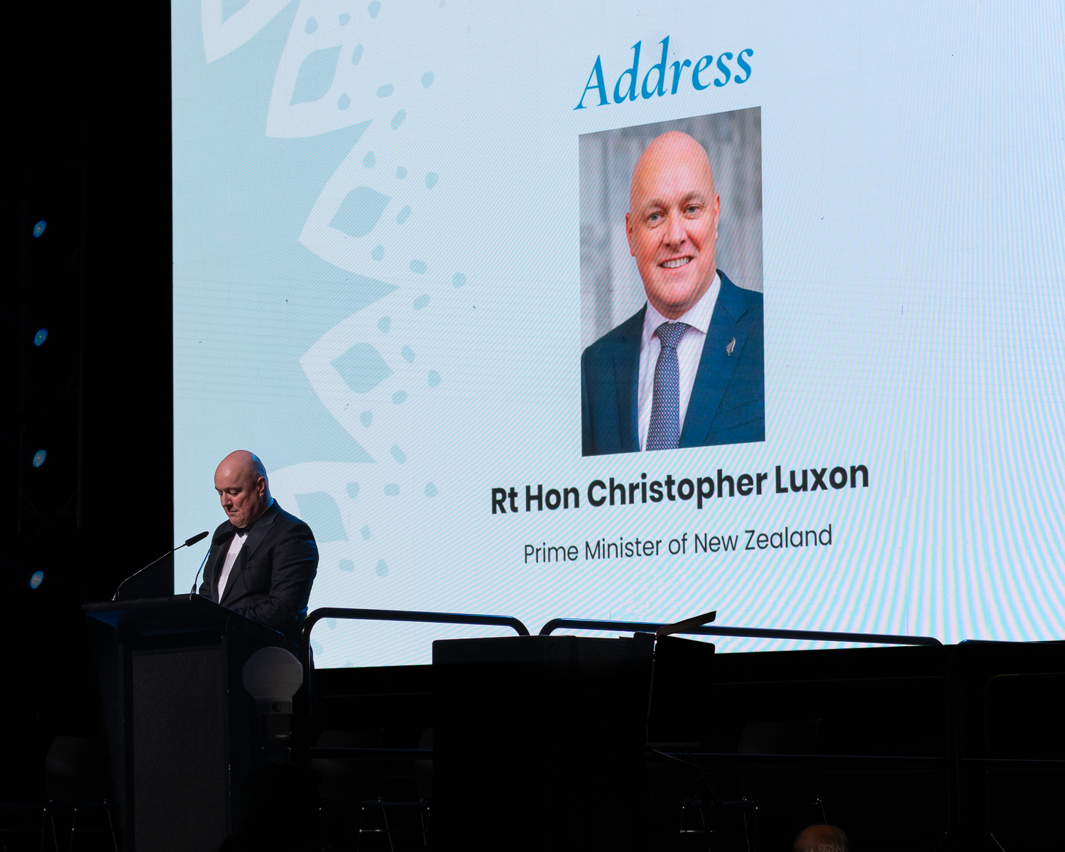 Speaker at podium beneath a projected screen showing Rt Hon Christopher Luxon, Prime Minister of New Zealand.
