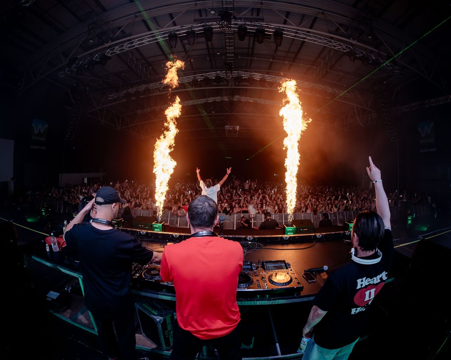 Three DJs perform on stage with dramatic fire columns shooting up behind them at a packed music festival.
