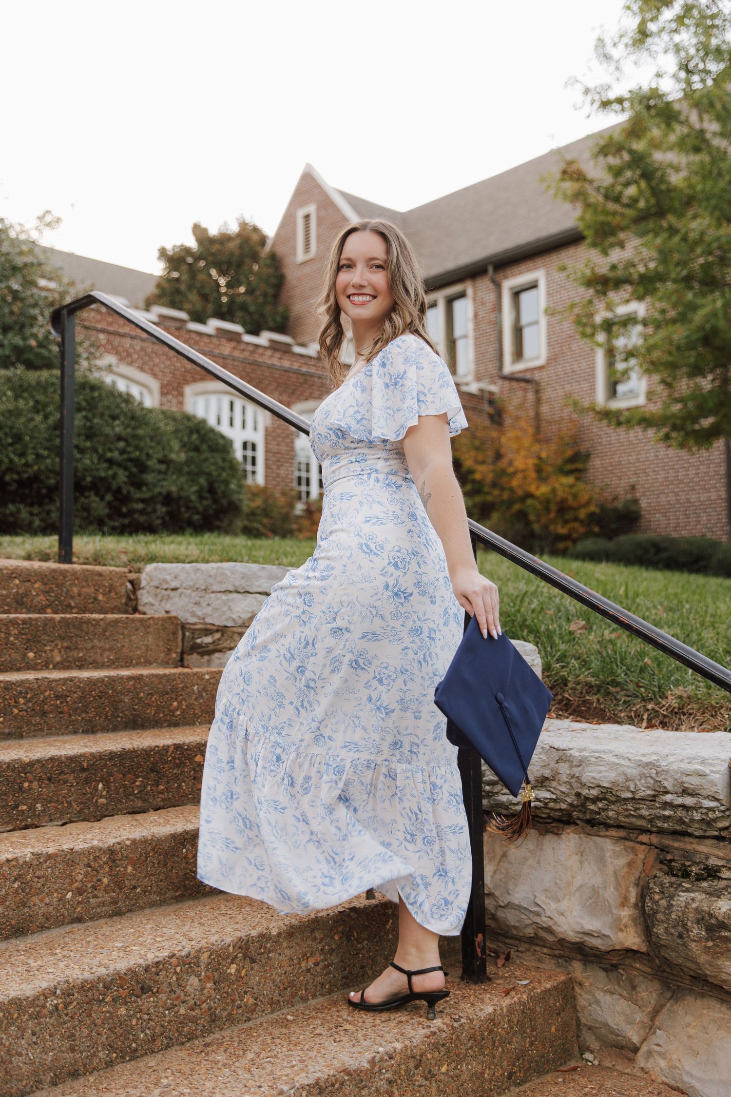 Graduate in floral dress sits gracefully on stone steps during outdoor photo session.