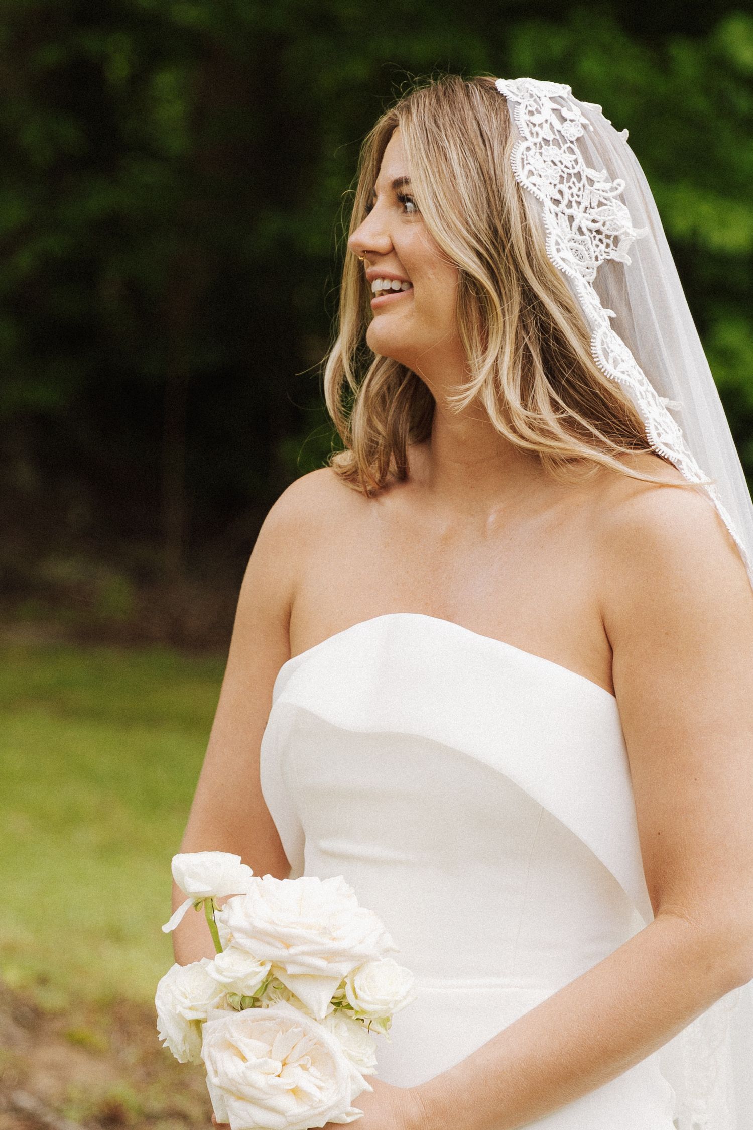 Bride in white strapless dress holding bouquet and smiling outdoors.